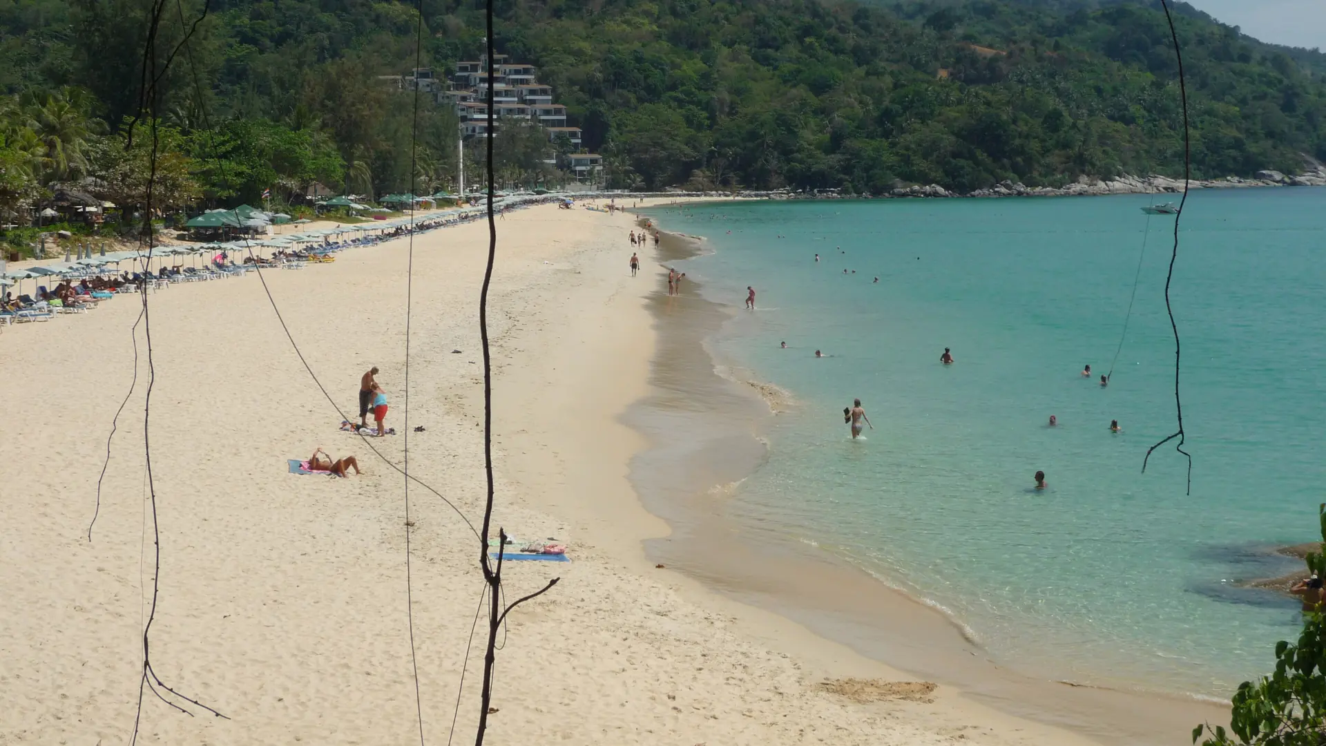 Adult women and other beachgoers beside turquoise shallows and a curving sandy beach on sunny Phuket