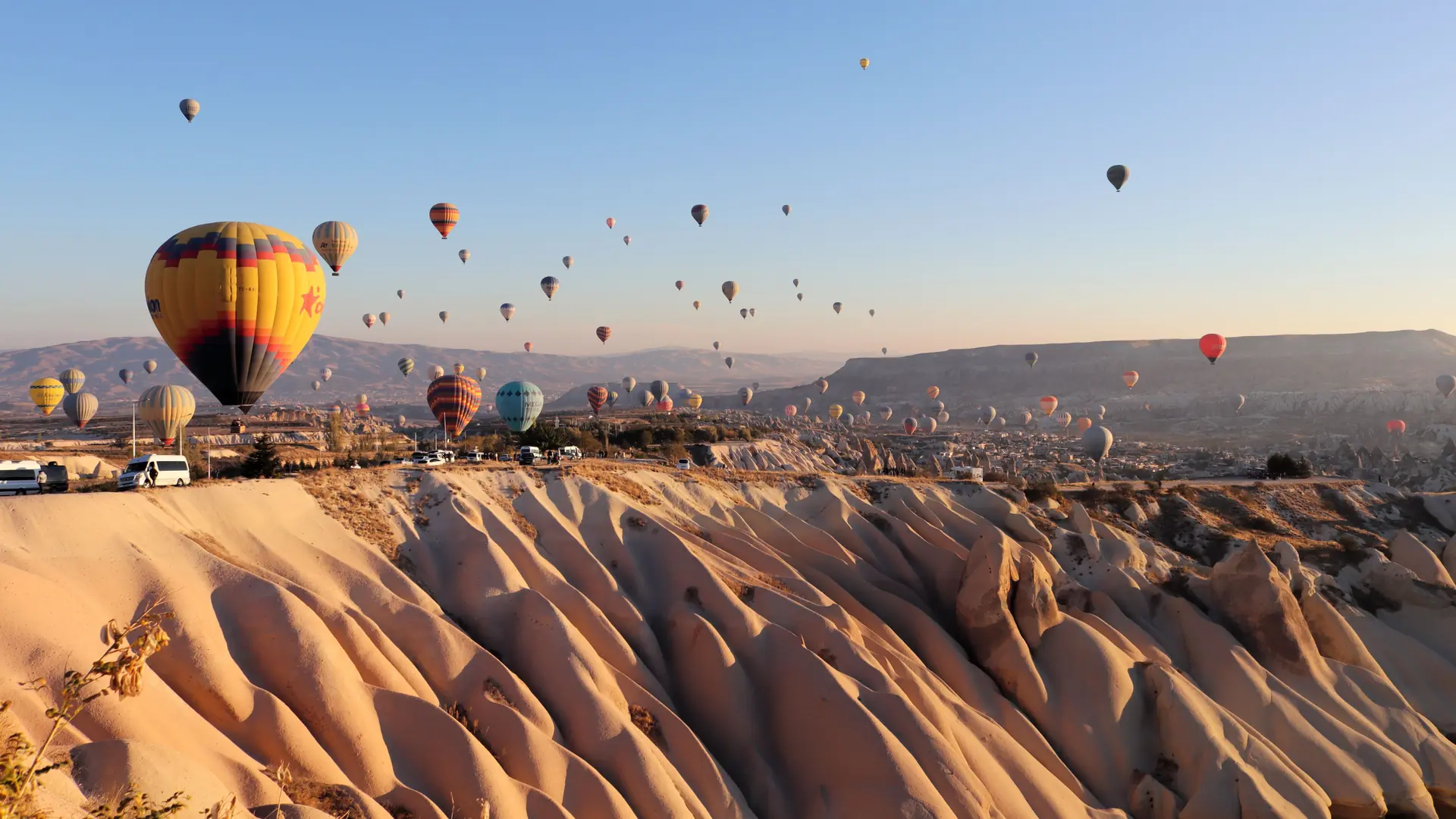Hot-air balloons over sunlit folded valleys in Cappadocia beneath a clear blue sky