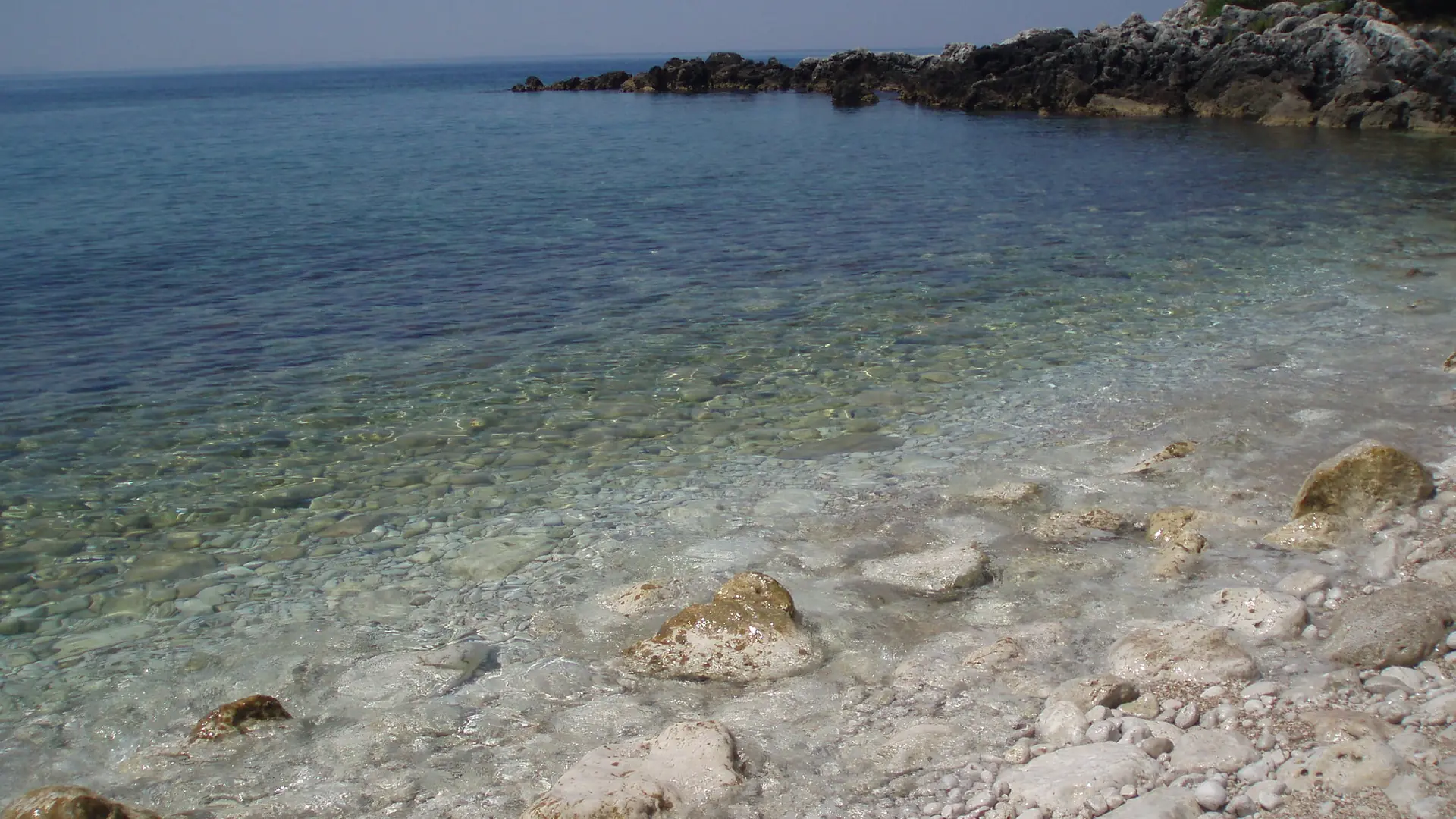 Clear water over the pebbles of a small cove at Himarë below a rocky headland on a sunny day