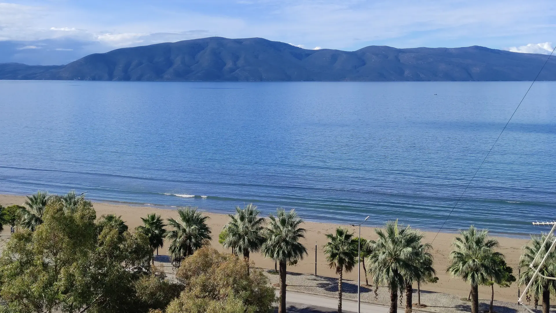 The curving bay of Vlorë with the Lungomare promenade, beaches and the Karaburun peninsula across calm blue water on a sunny day