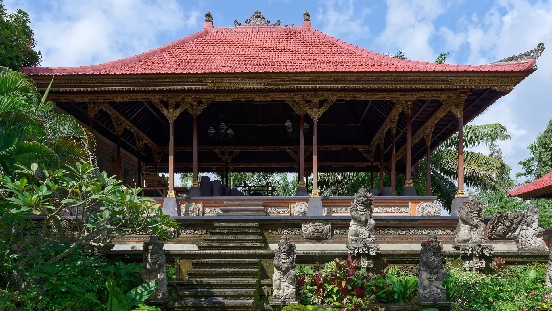 Carved stone guardians and a red-roofed pavilion at Ubud Palace beneath a blue sky