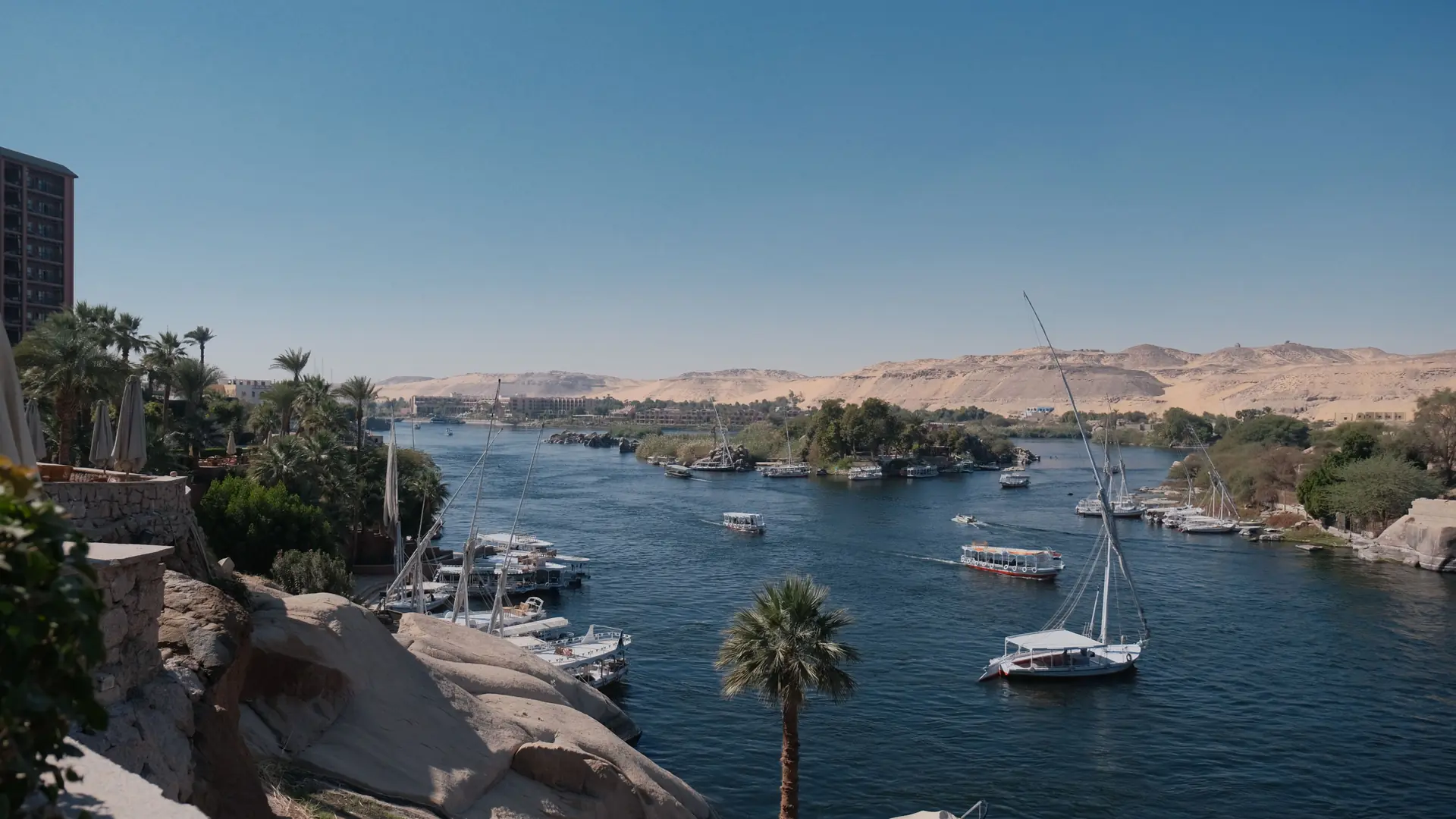 Boats on the Nile beside Aswan islands and desert hills in bright daylight.