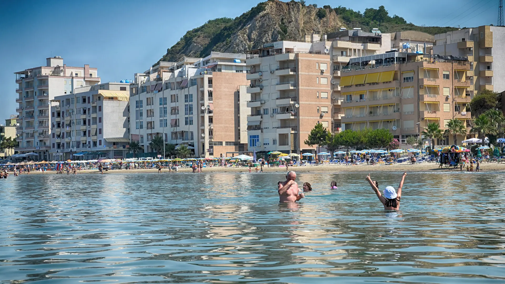 Swimmers below apartment blocks and the rocky headland of Shkëmbi i Kavajës on the Durrës beach strip on a sunny day