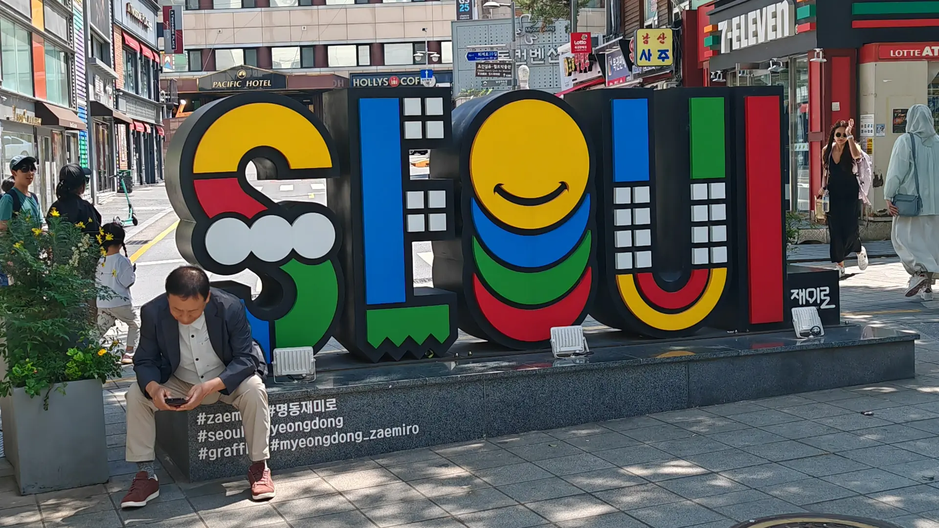 A large, colourful 'SEOUL' sculpture sign on a Myeongdong street corner in bright daylight, with 7-Eleven and hotel signage behind it
