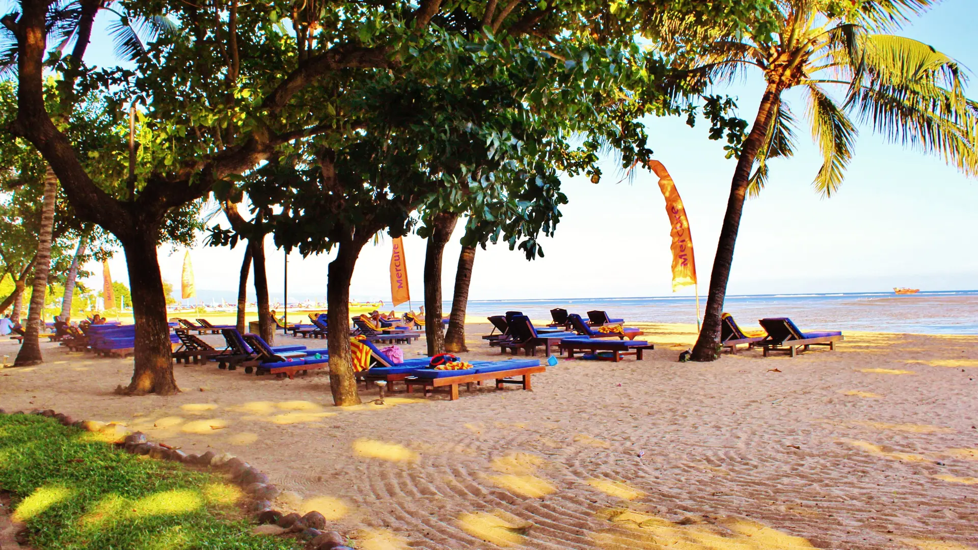Sun loungers beneath palms and shade trees beside Sanur beach in Bali