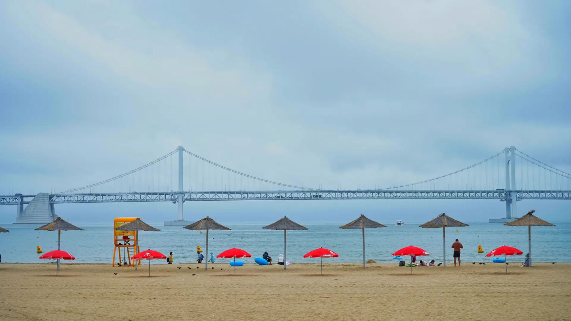 Gwangalli Beach with straw umbrellas and a lifeguard stand, the Gwangan Bridge spanning the water behind, in bright daylight