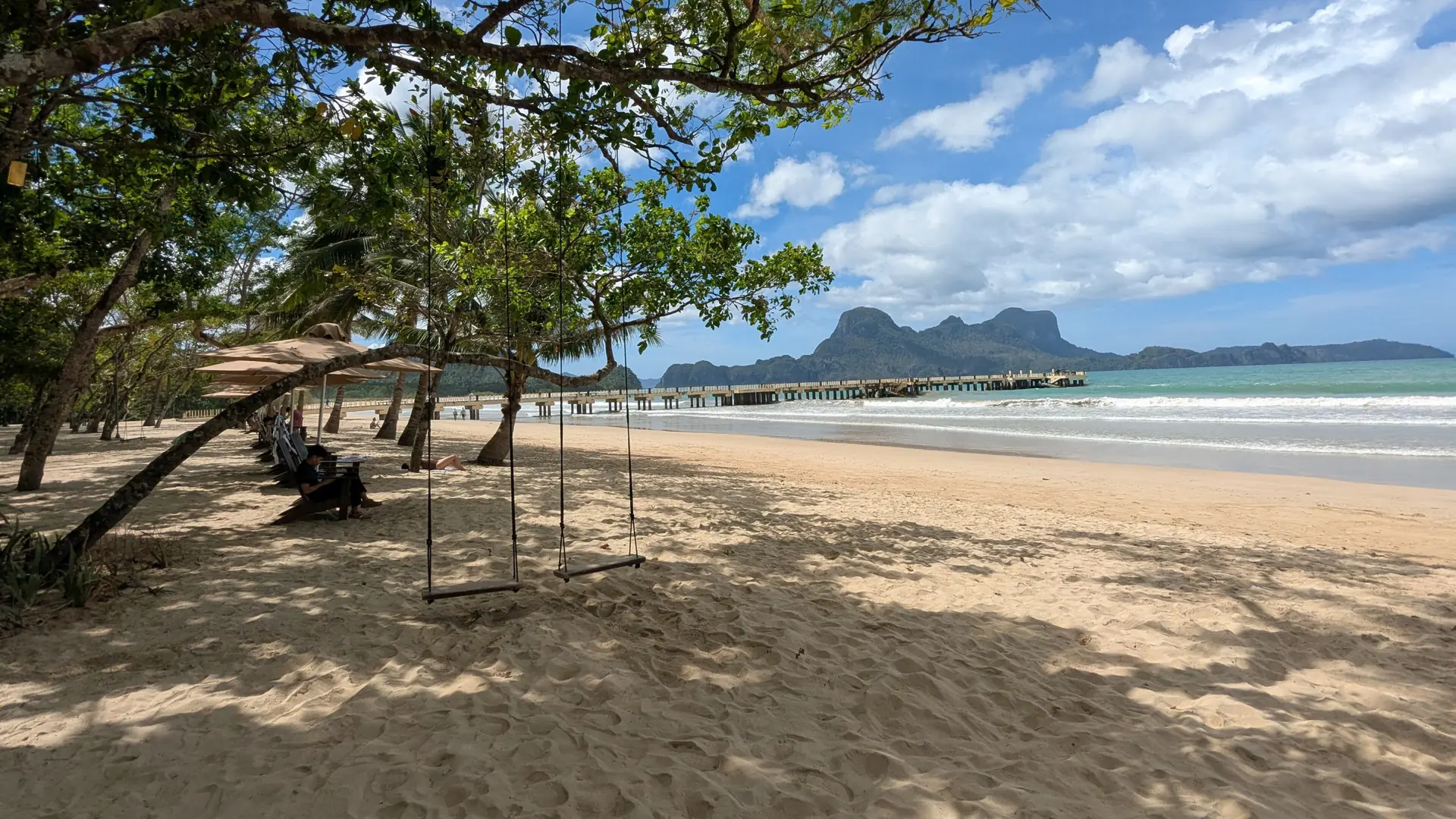 Wooden swings beneath trees at Lio Beach, El Nido, beside breaking waves and a pier