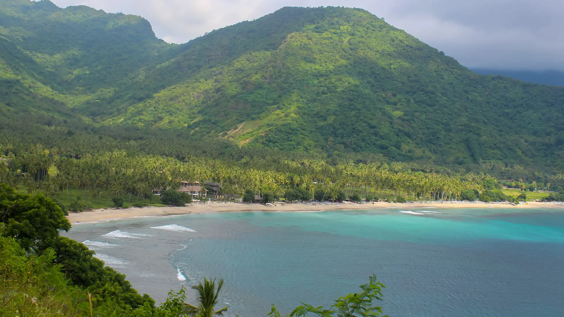 Palm-lined beach and green hills around a turquoise bay in the Senggigi coastal region of western Lombok