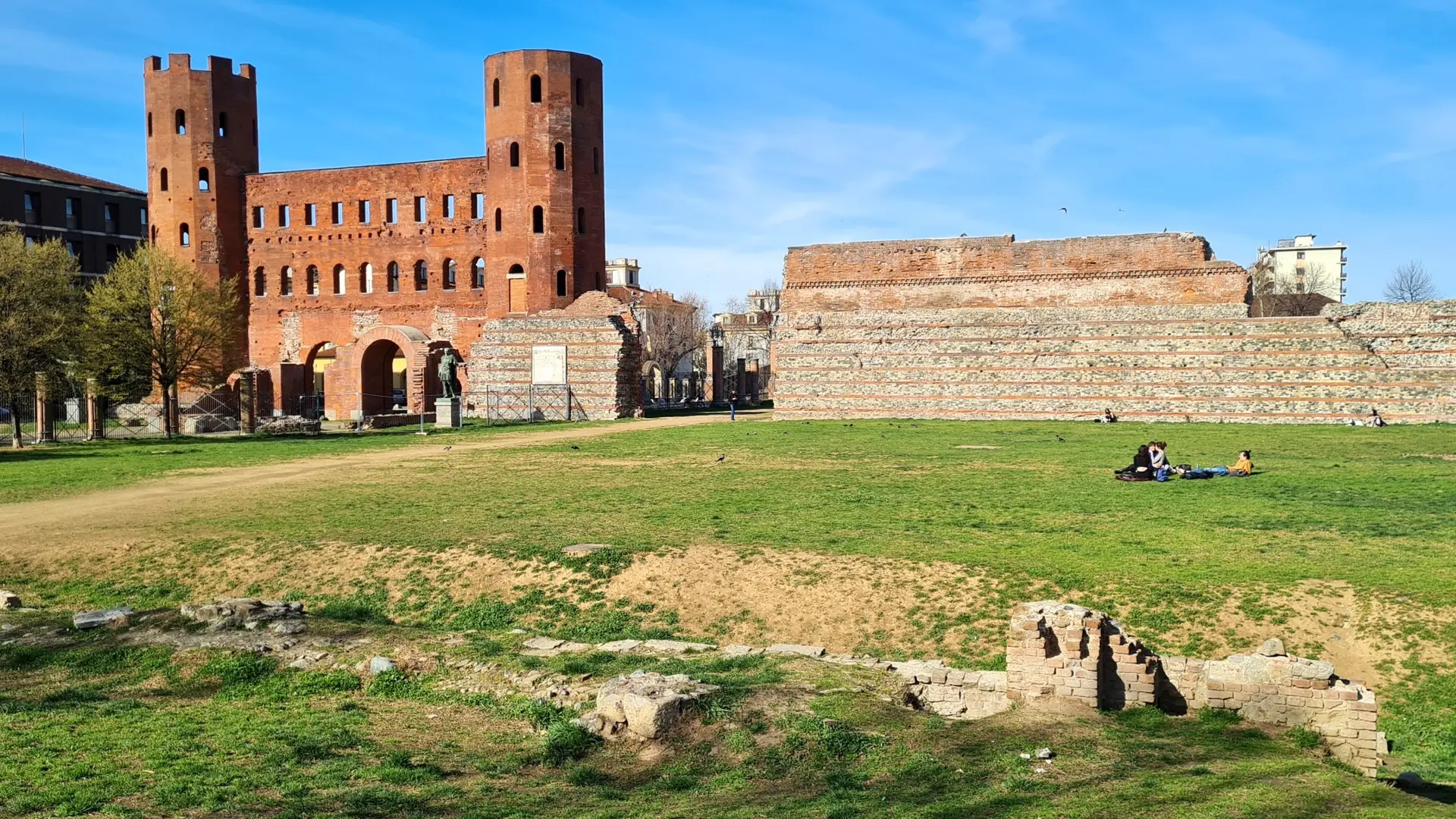 Porta Palatina’s brick towers and arched gateways above archaeological remains and a sunlit lawn.