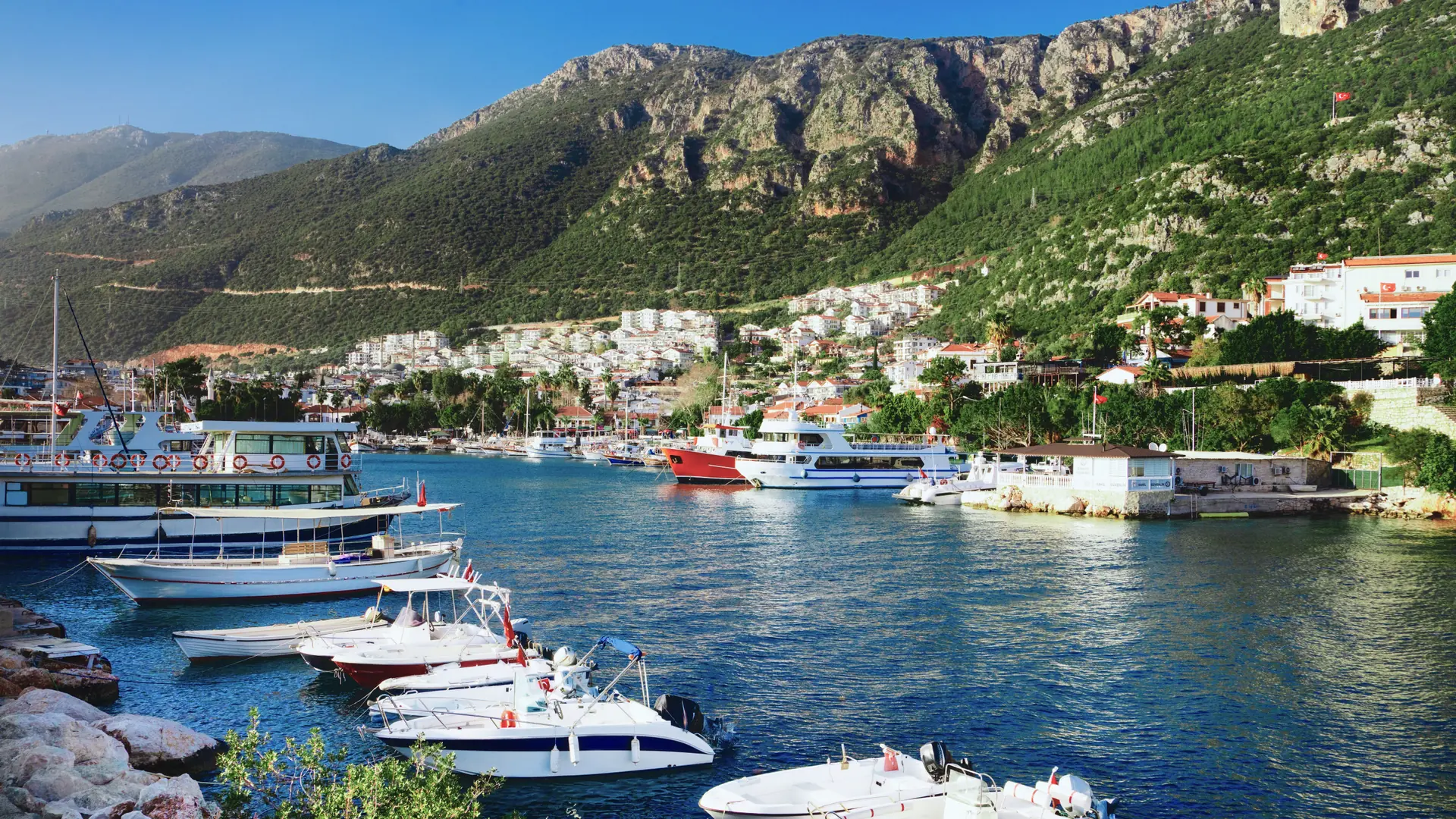 Sunlit boats in Kaş old harbour below white hillside homes and a limestone cliff