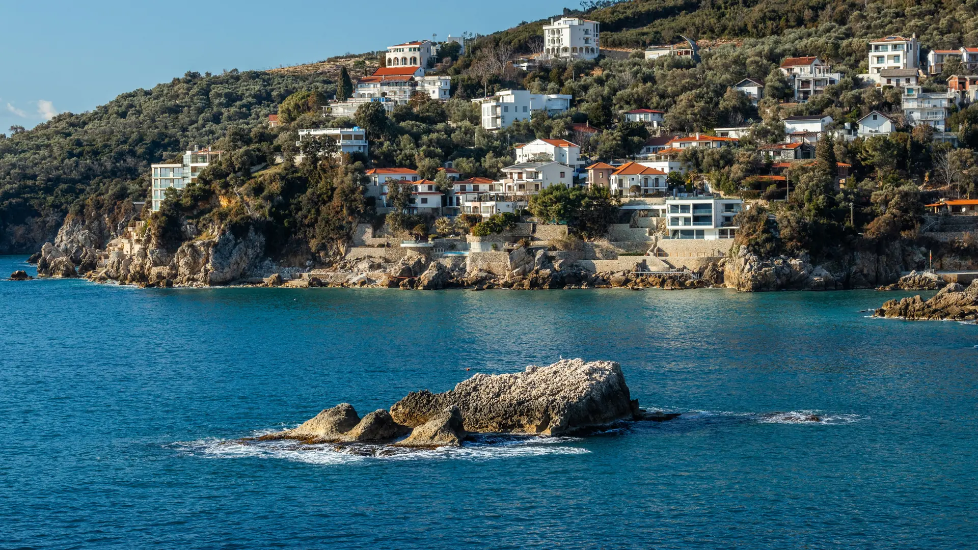 Rocky Ulcinj coastline below hillside houses, seen across clear blue water