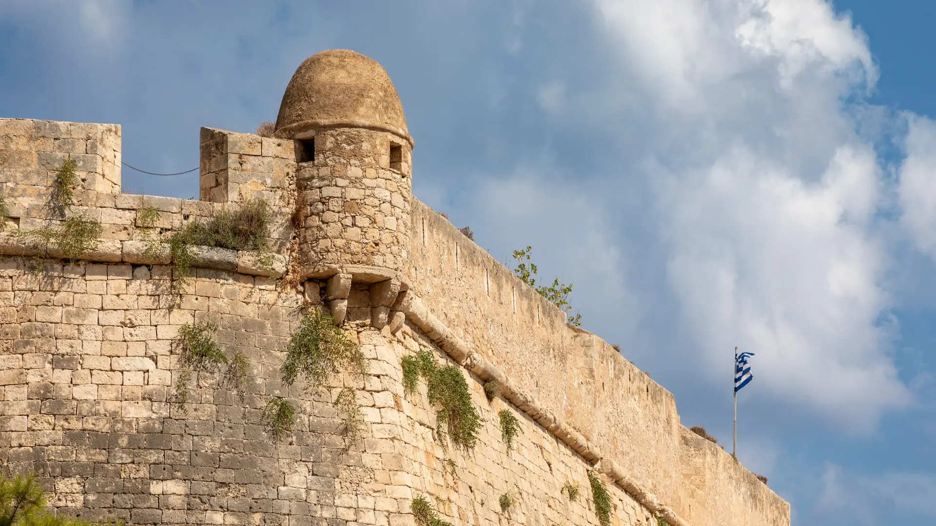 Sunlit stone walls and a projecting turret of the Fortezza fortress in Rethymno beneath a blue, cloud-filled sky.