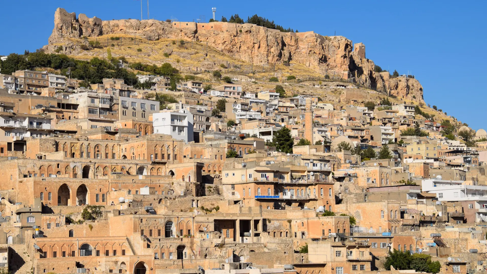 Sunlit Mardin stone houses rising toward the flat-topped citadel beneath a clear blue sky