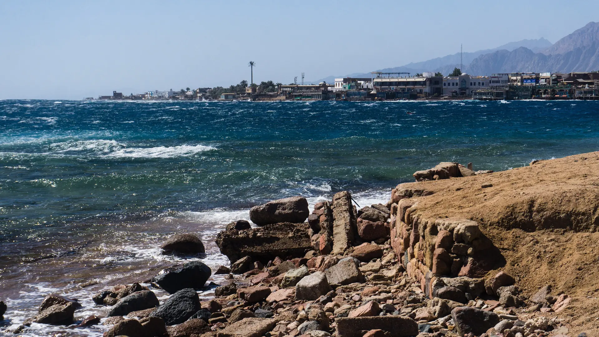 Sunlit rocky shore at Lighthouse Reef in Dahab’s Masbat Bay, with waves and waterfront buildings