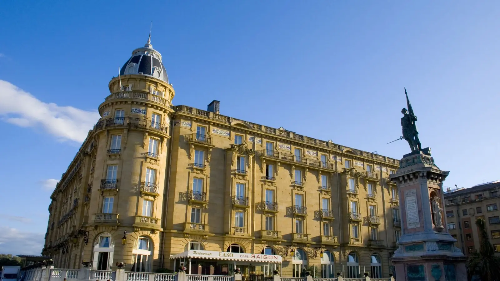 Hotel María Cristina’s sunlit façade and domed corner, with a bronze monument beside the Urumea