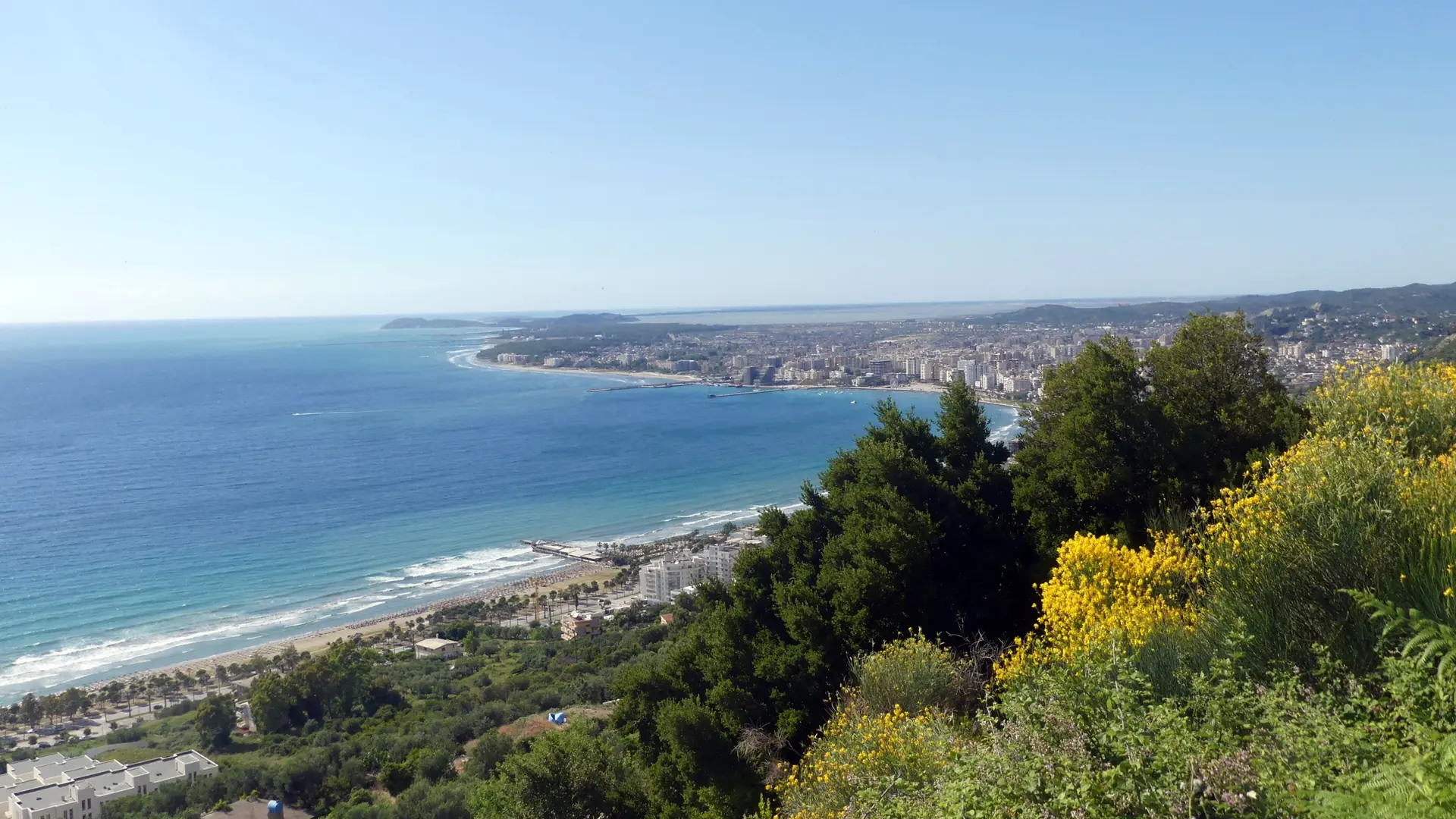 The curve of the Bay of Vlorë and the city's beaches seen from a green hillside on a sunny day
