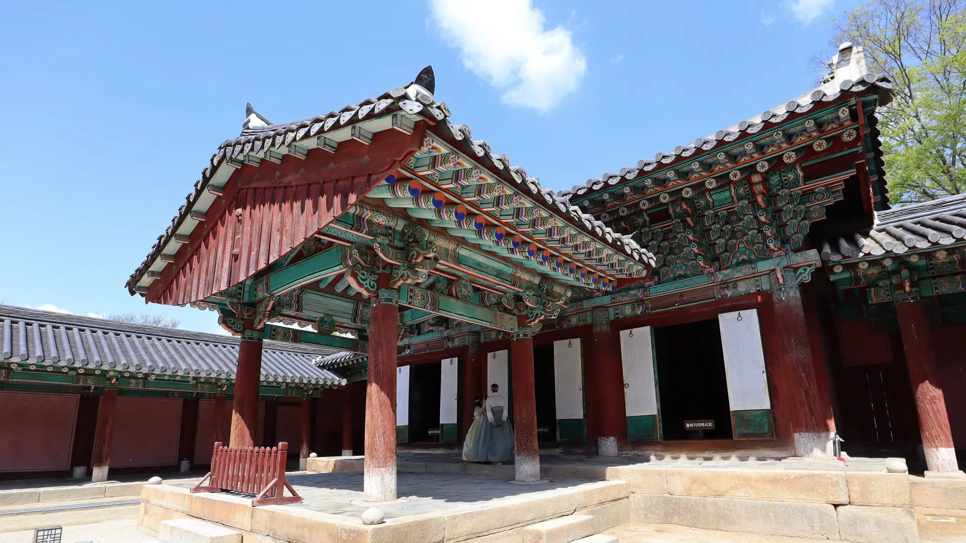 Painted wooden halls and broad eaves at Gyeonggijeon Shrine in Jeonju, with a visitor in the courtyard
