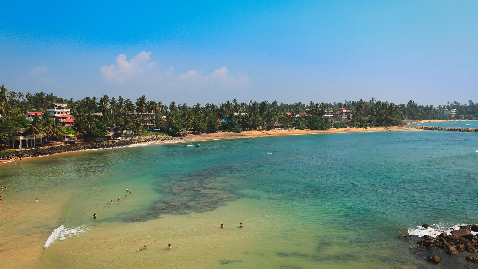 Aerial view across the shallow turquoise water and palm-lined sand of Mirissa Beach