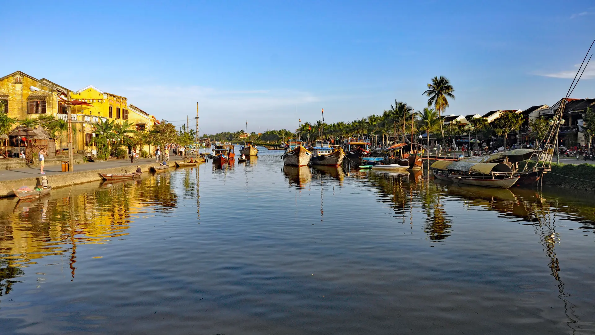 Boats on the Thu Bon River beside Hoi An heritage houses under a blue sky