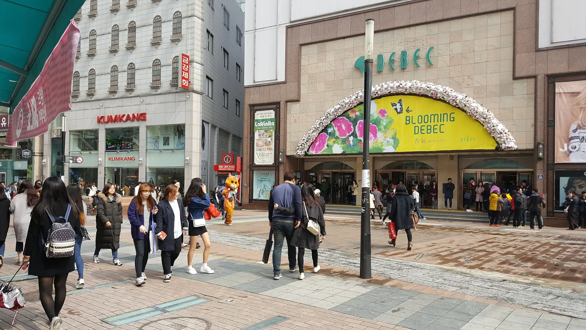 A busy Dongseongno street in Daegu, crowds passing a department store with a floral banner arch, in bright daylight