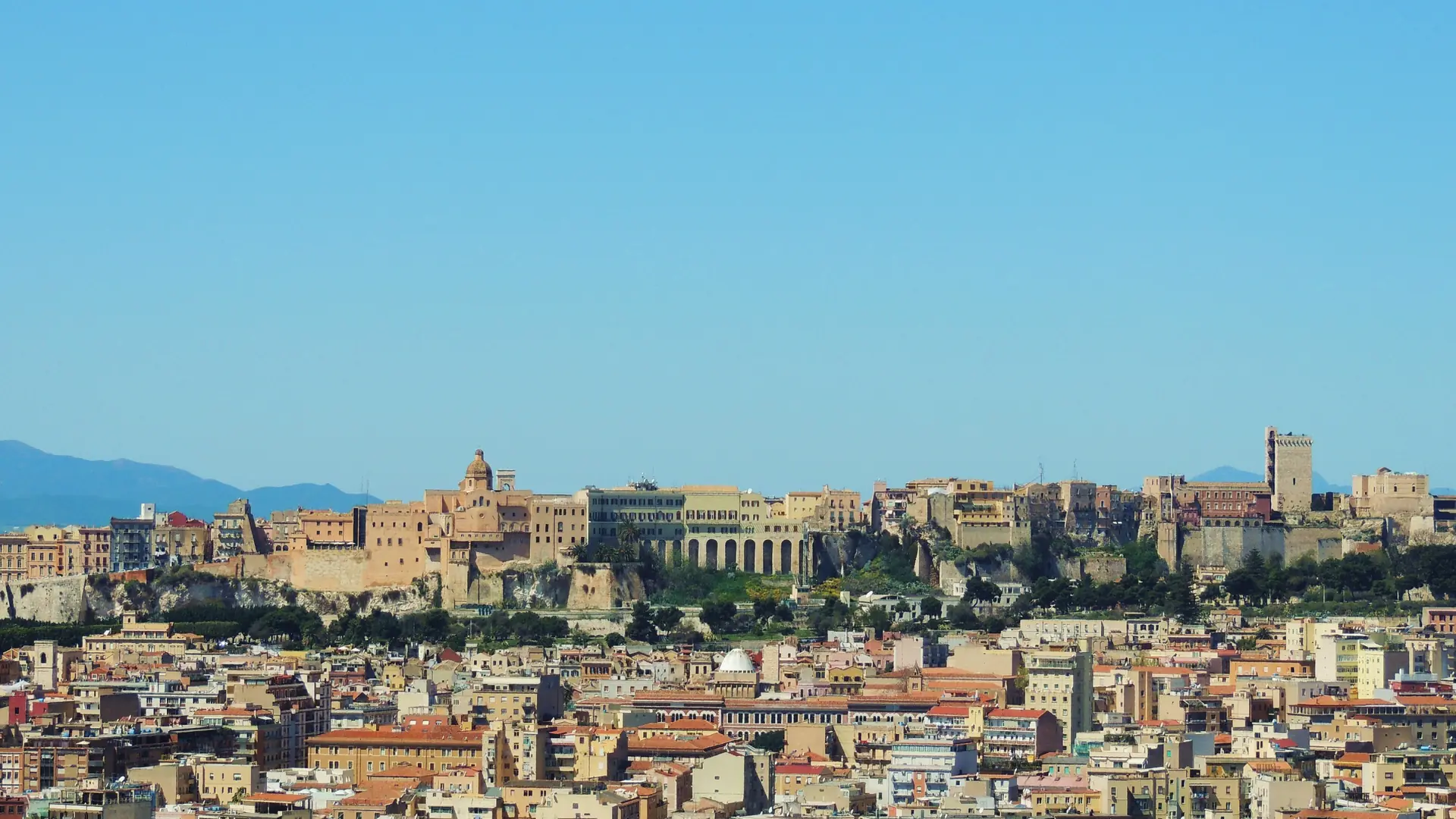Cagliari’s Castello quarter, cathedral dome and stone towers above the city’s rooftops under a clear blue sky.