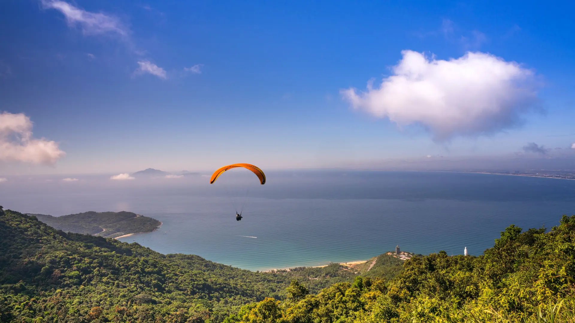 An orange paraglider above forested coastal hills and a blue bay in Da Nang