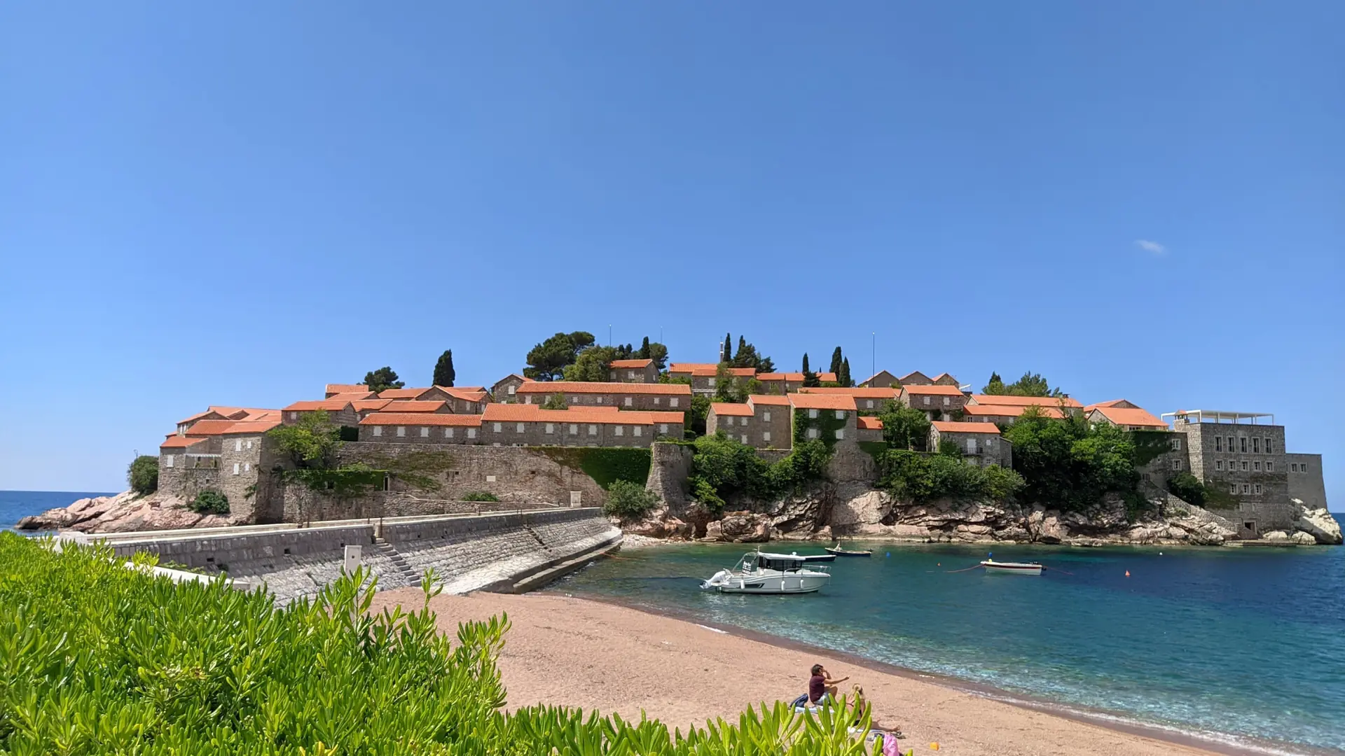 Fortified Sveti Stefan island, its causeway and a sheltered shore in bright daylight