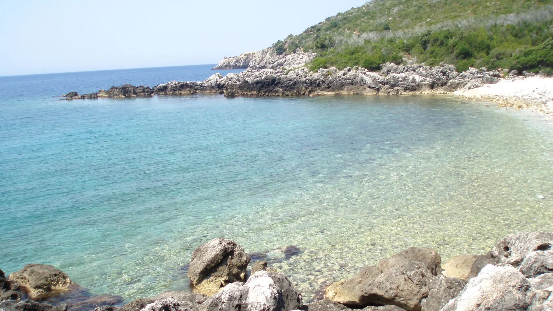 Clear shallow water and a pebble shore below a green headland at Himarë on a sunny day