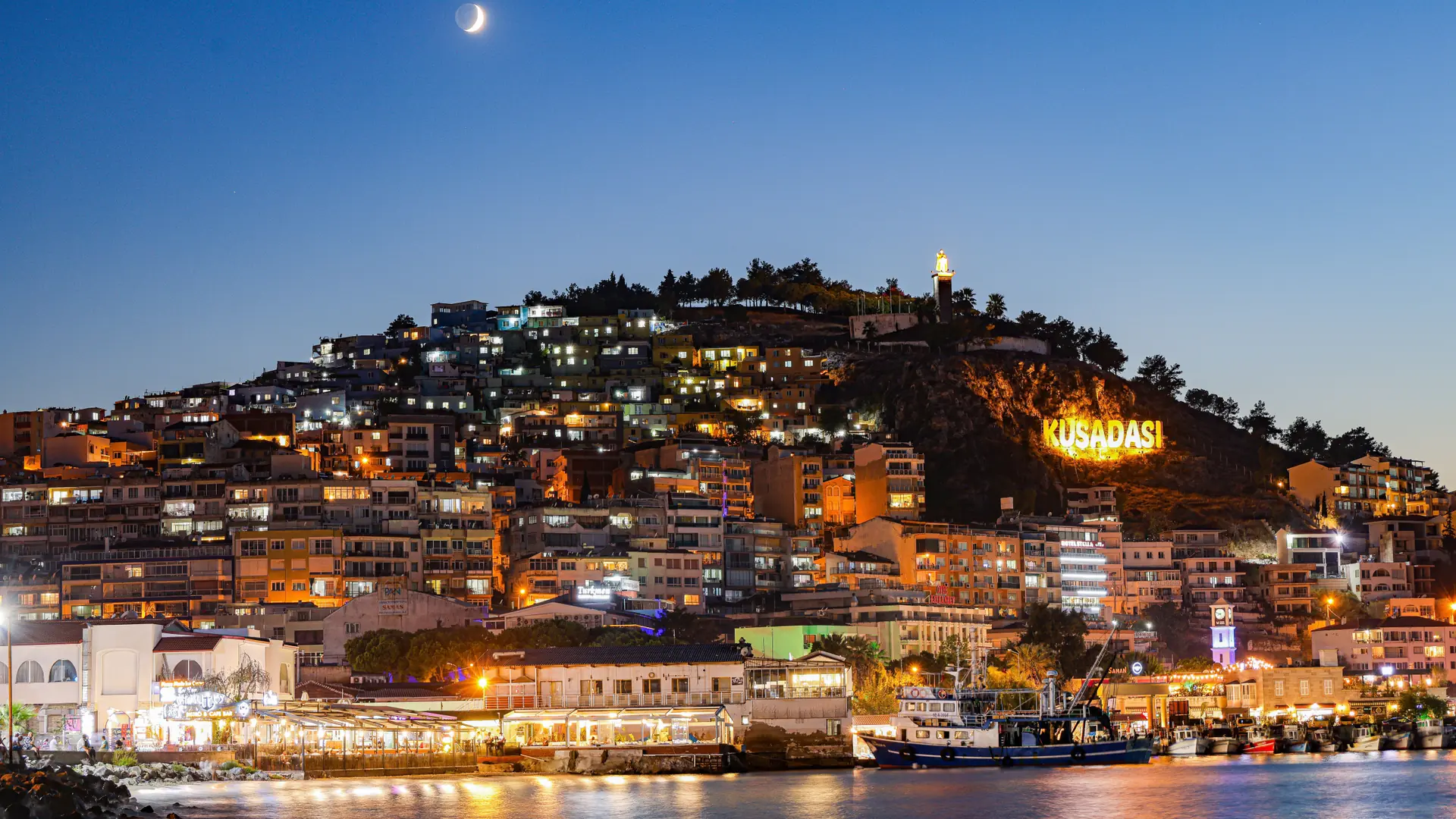 Kuşadası waterfront lights reflected across the harbour beneath a crescent moon
