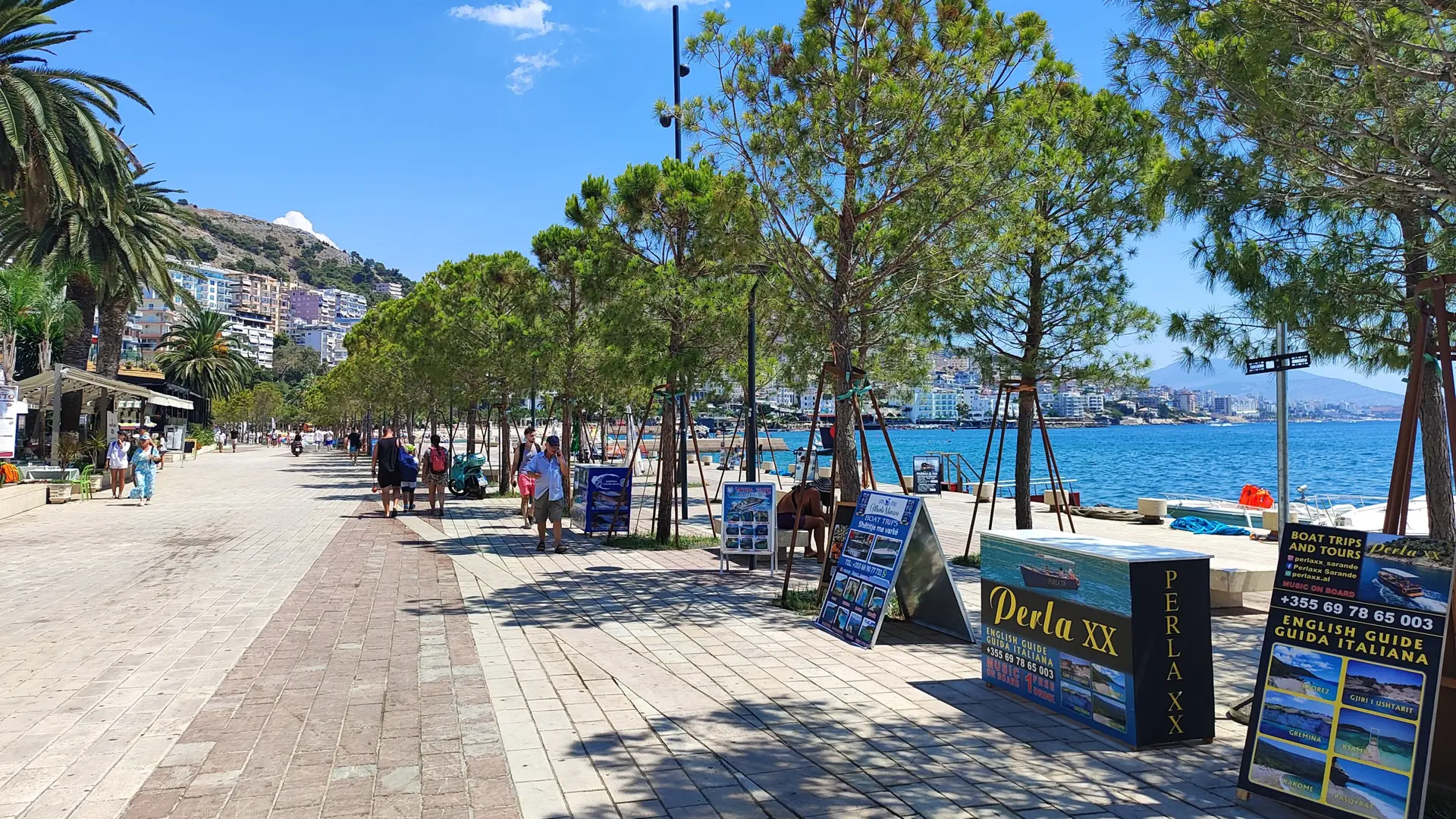 The tree-shaded seafront promenade in Sarandë, with tour boards and the bay beyond, on a sunny day