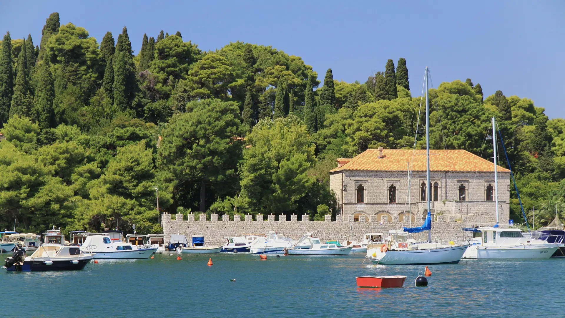 A stone-walled marina near Dubrovnik with moored boats, pines and a tiled-roof villa