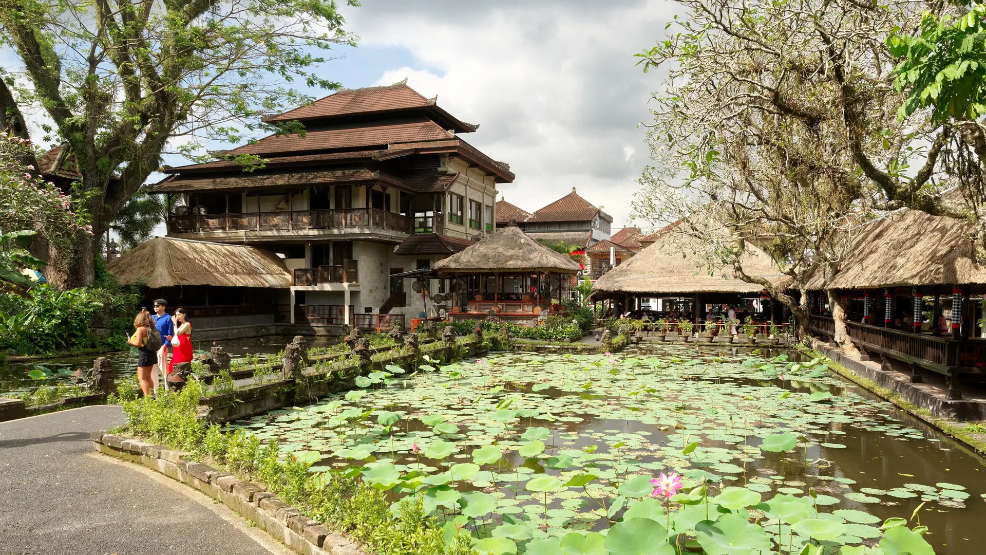 Sunlit lotus pond beside Balinese buildings and thatched pavilions at Pura Taman Saraswati in central Ubud