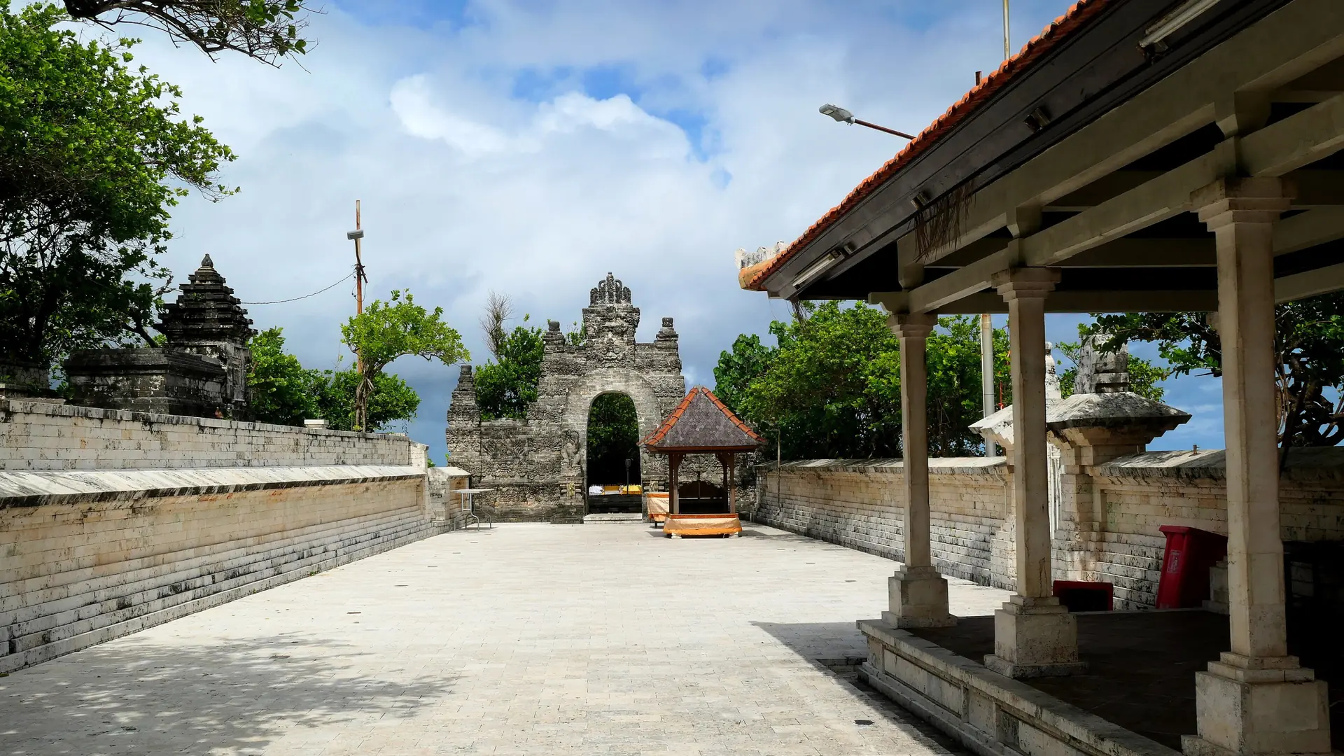 Sunlit courtyard, carved stone gateway and red-roofed pavilion at Pura Uluwatu in wider Pecatu, Bali