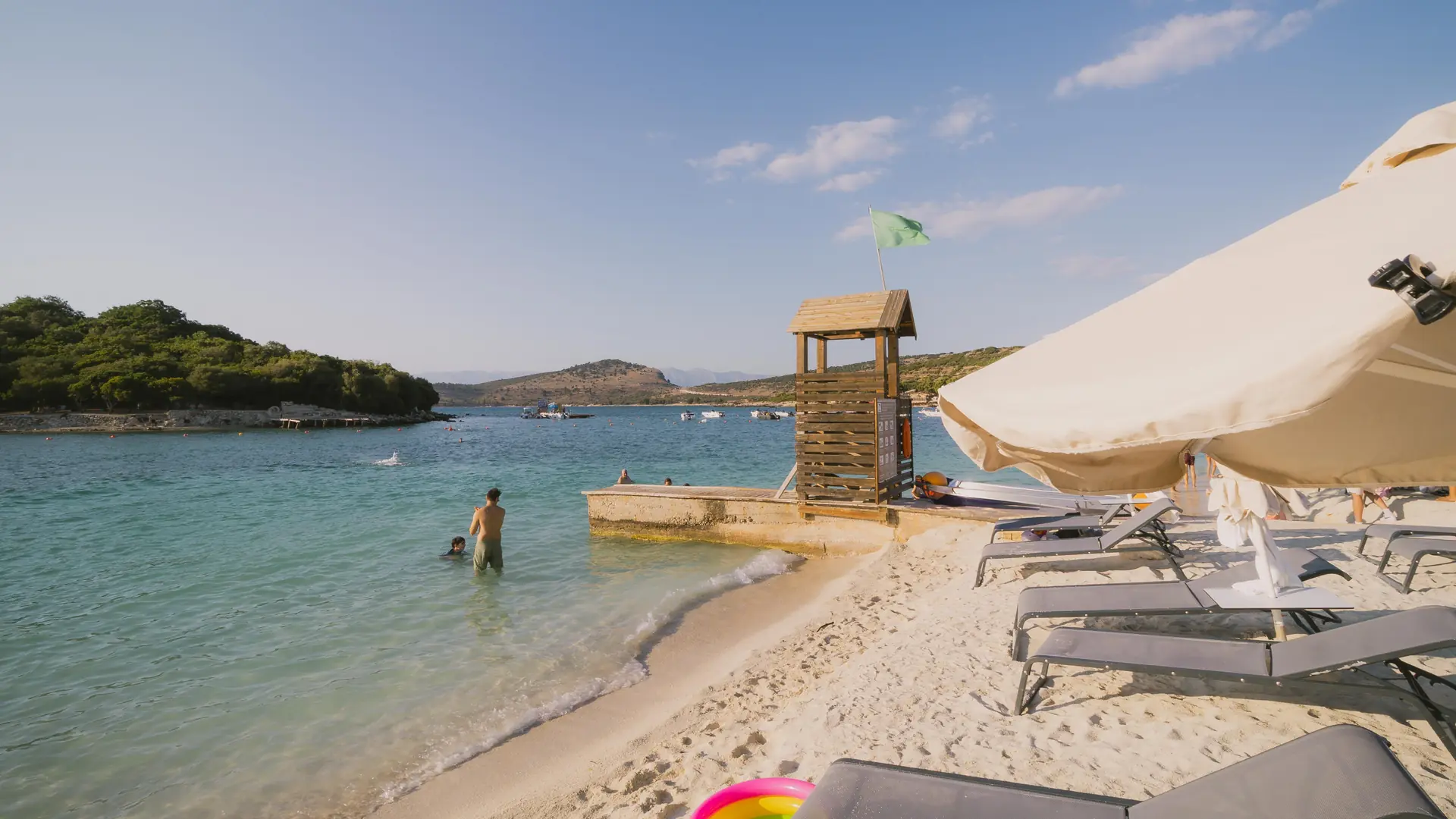 The wooded Ksamil islets across a channel of clear turquoise water seen from the white-sand beach at Ksamil on a sunny day
