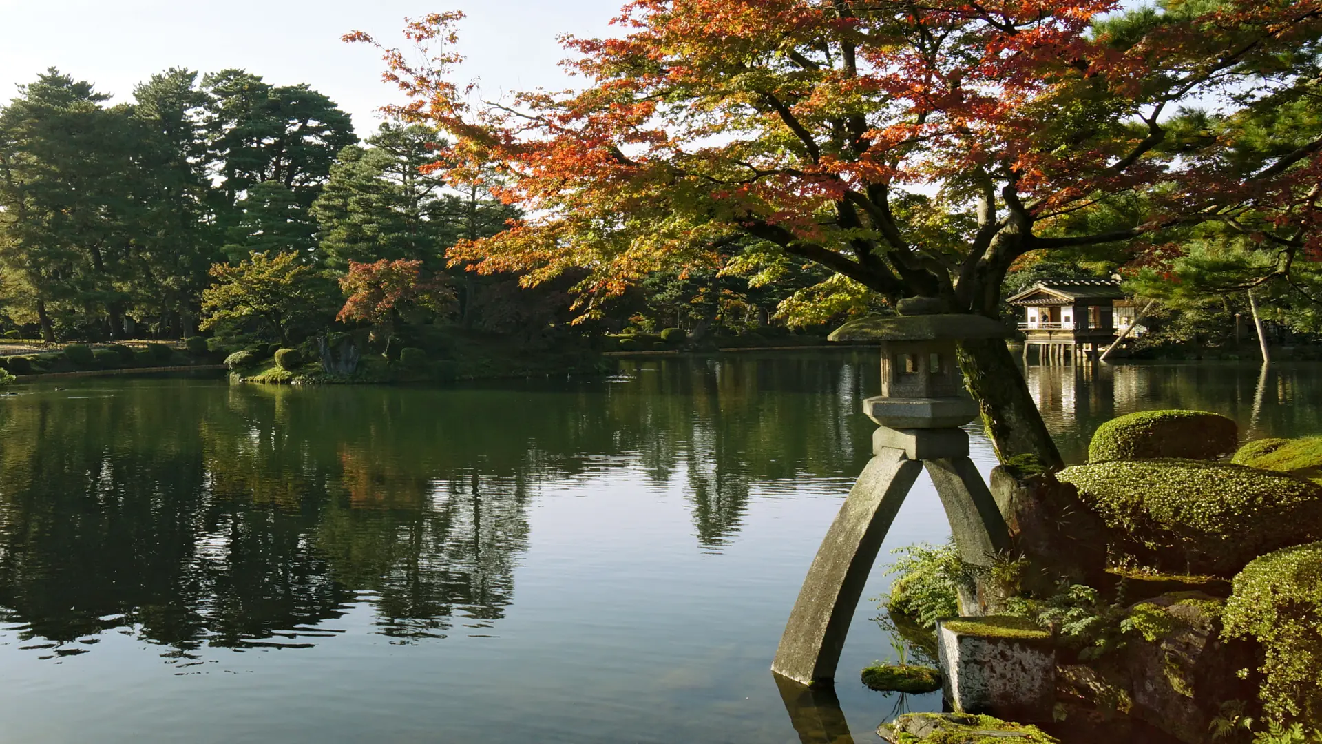 Kotoji lantern beside Kasumigaike Pond and autumn trees in sunlit Kenrokuen.