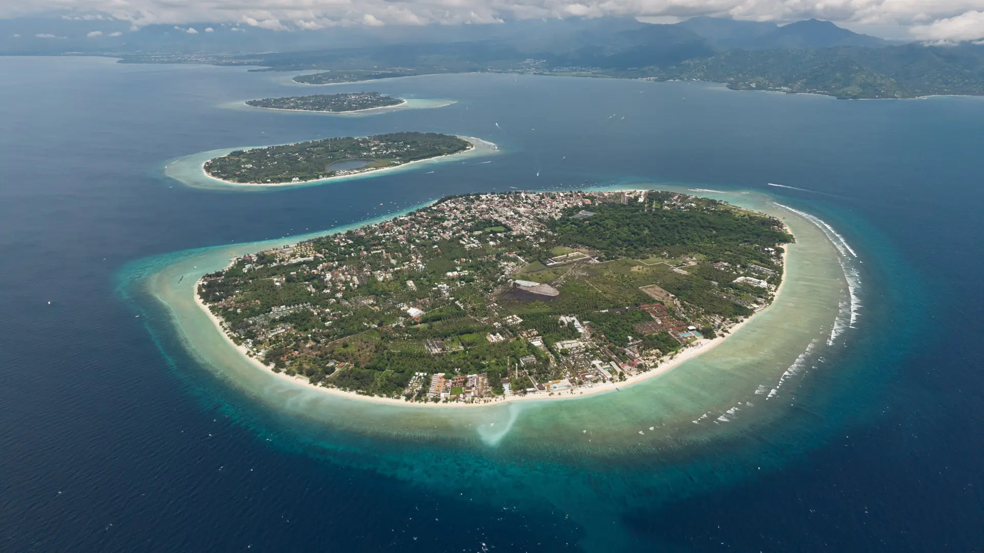Wider aerial view of Gili Trawangan in March 2026, with green interior, sandy coast and neighbouring islands