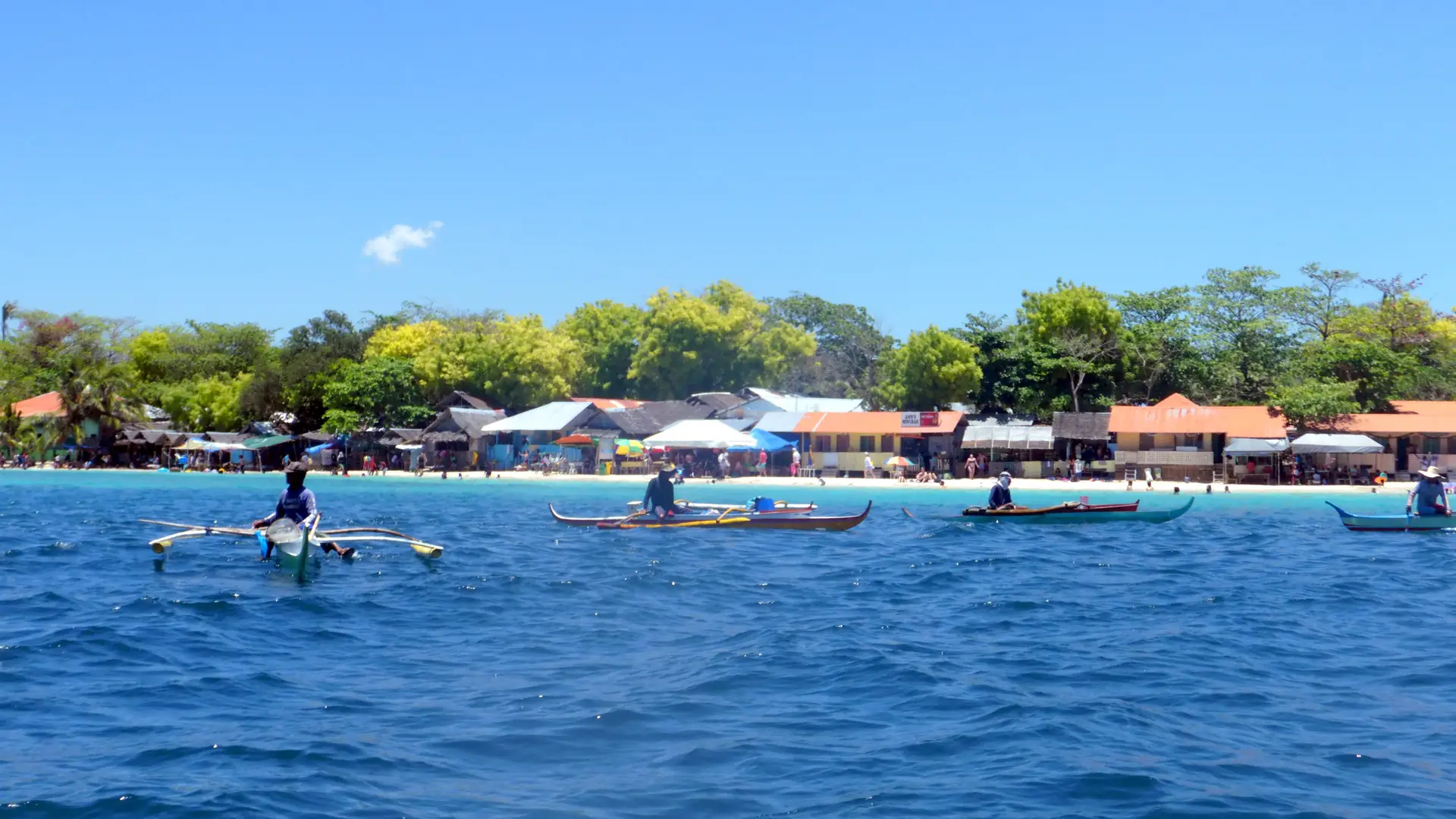 Outrigger boats beside Moalboal’s sandy shoreline, beach buildings and trees under a blue sky