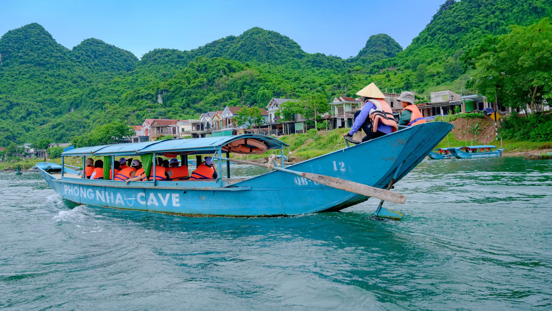 A blue Phong Nha Cave passenger boat on the Son River beside village houses and green limestone hills