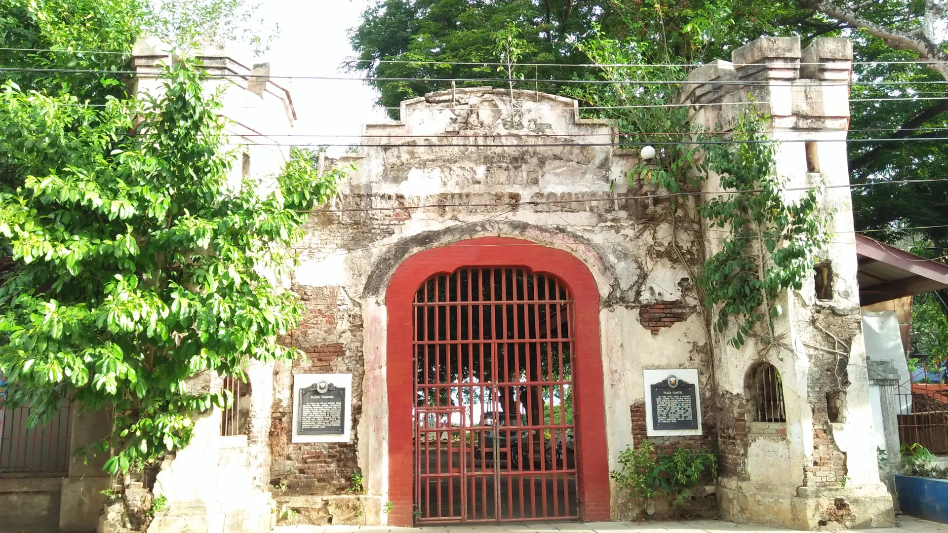 The red gate and stone entrance of Plaza Cuartel beneath green trees in Puerto Princesa