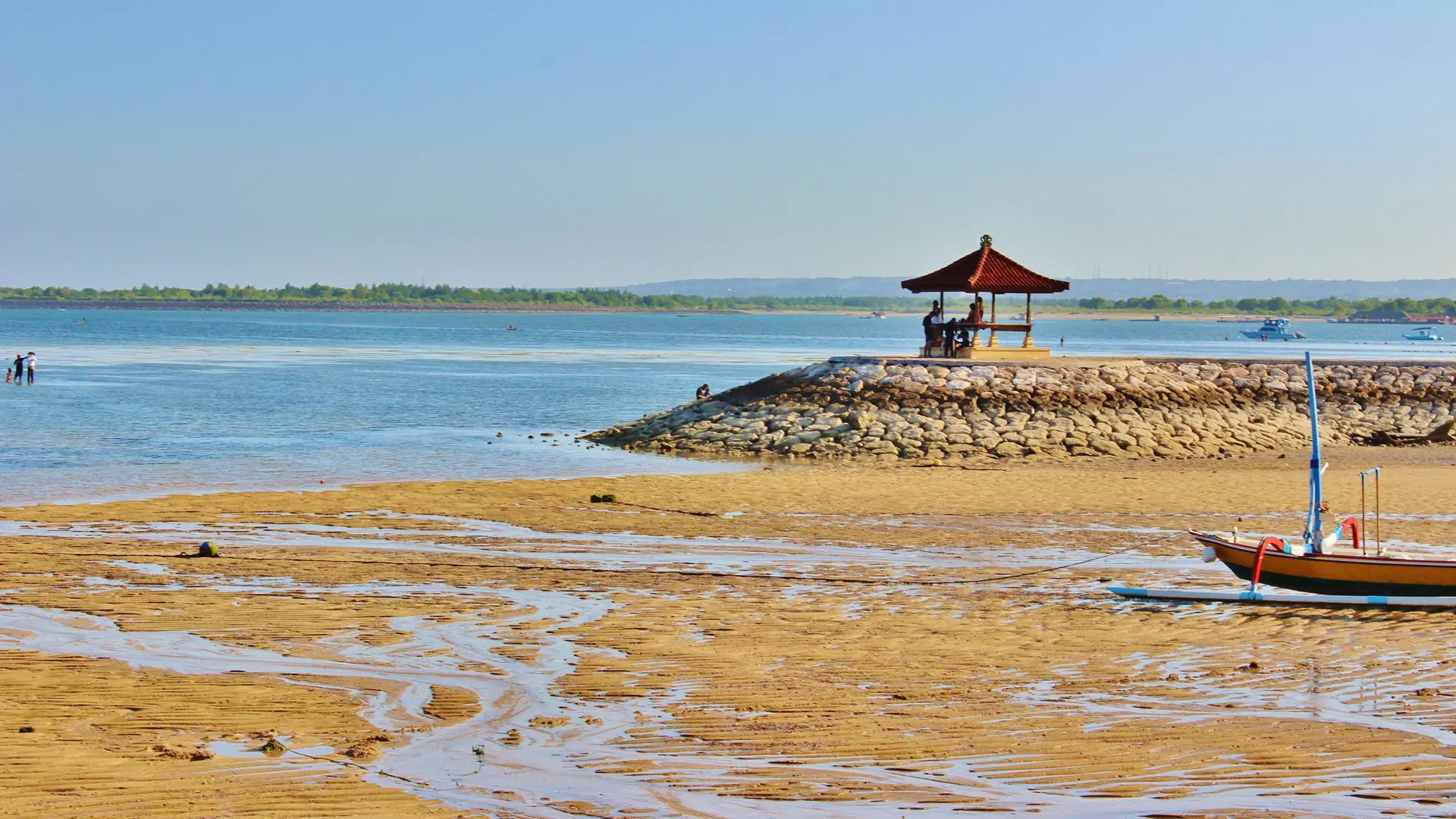 A red-roofed pavilion beside shallow blue water and exposed sand on Sanur Beach