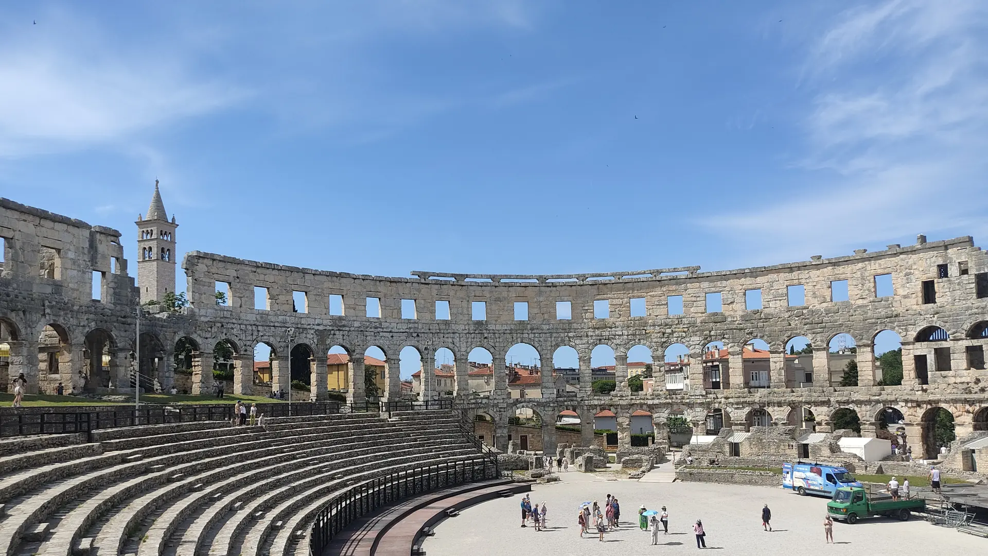 Inside Pula Arena's Roman amphitheatre under a bright blue sky, with the bell tower visible through the arches