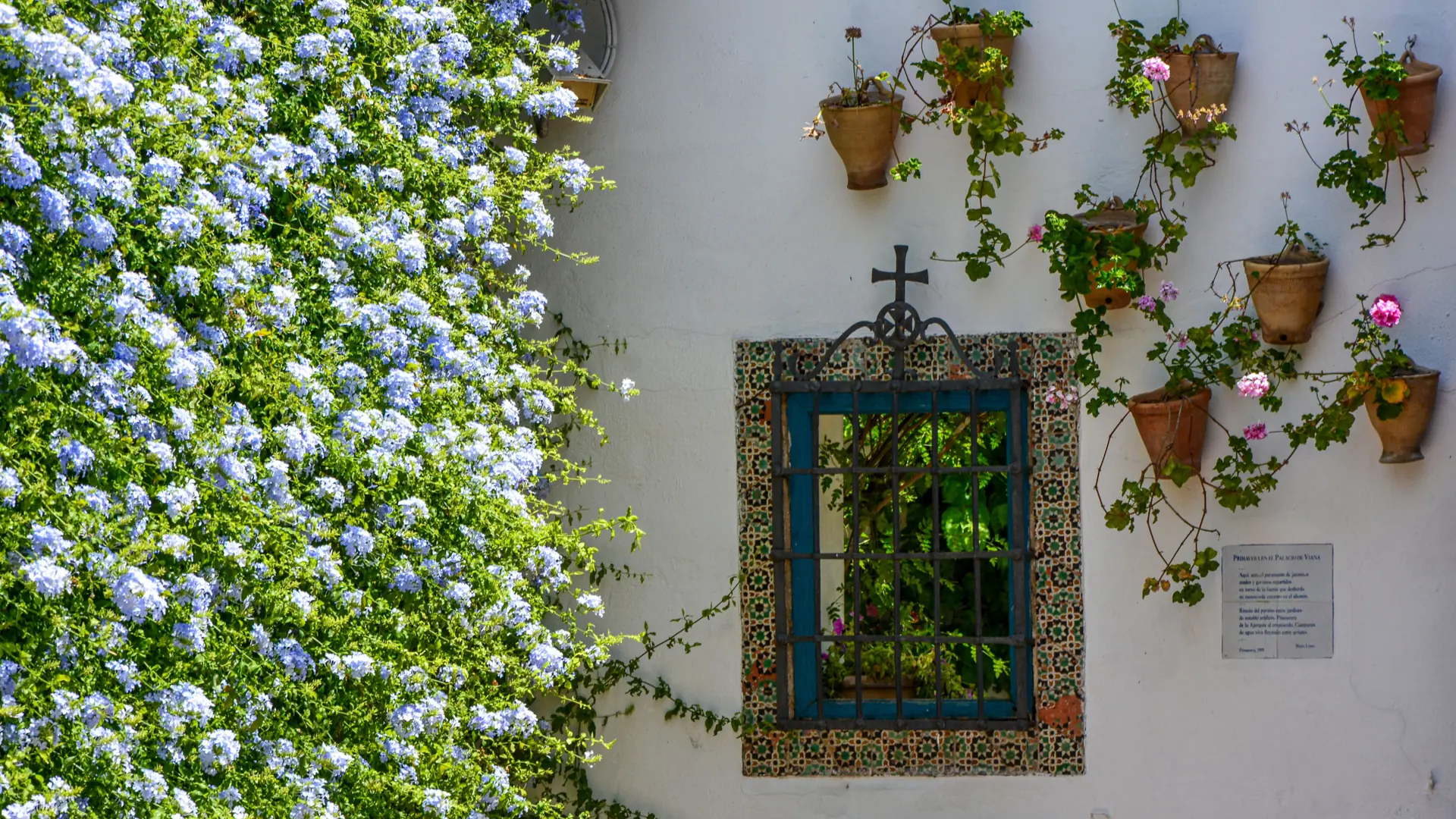 Blue tiled window and iron cross beneath hanging flowerpots in Palacio de Viana’s Patio de los Jardineros, Córdoba