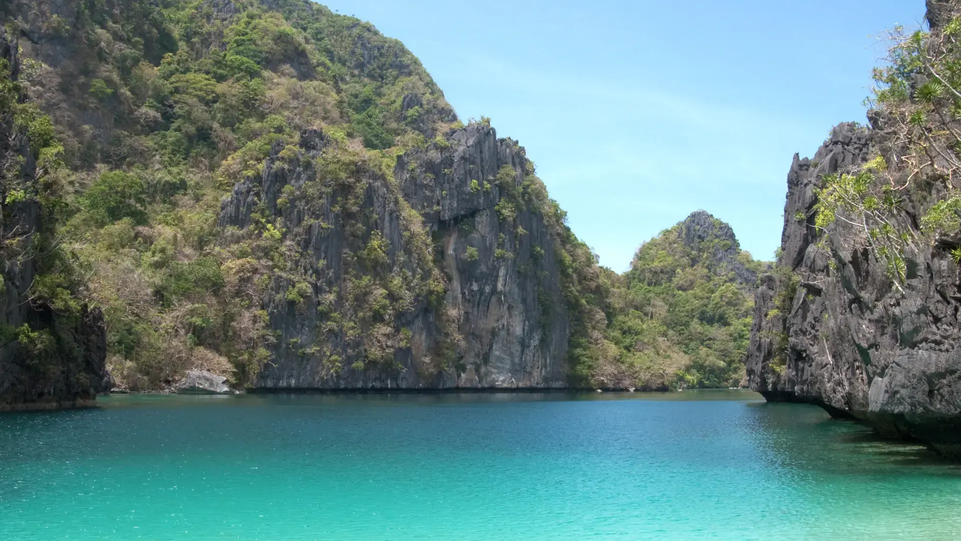 Sunlit limestone islands around blue-green water beneath a clear sky in Bacuit Bay, El Nido