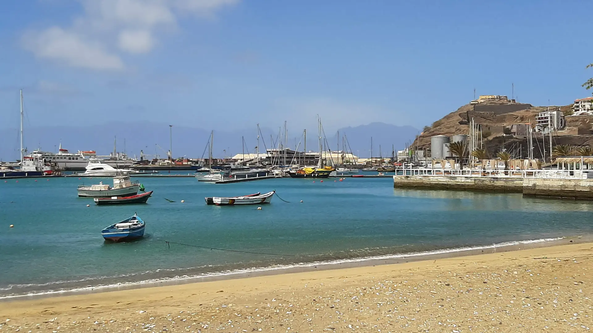 Sandy edge of Mindelo harbour with small boats and waterfront buildings in the sun
