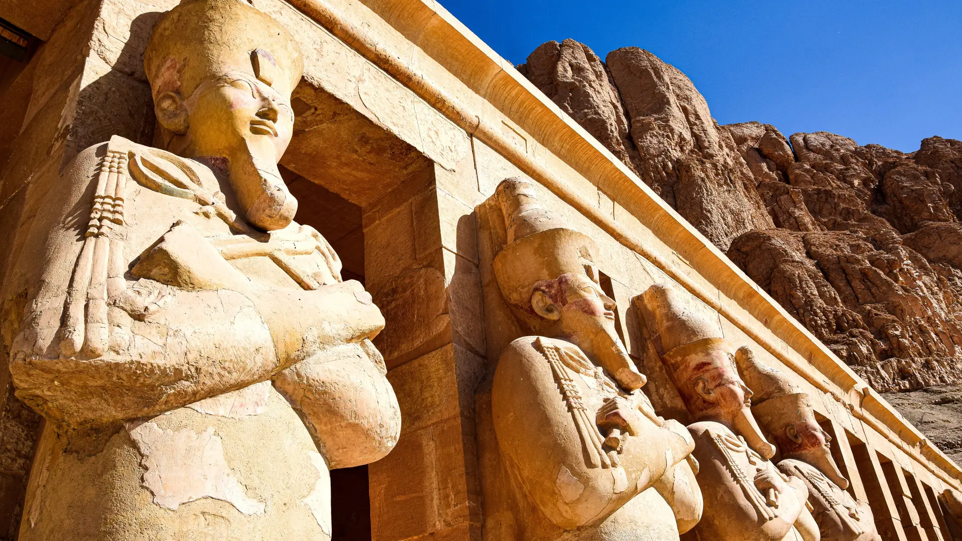 Sunlit statues of Hatshepsut at her Luxor temple against rocky cliffs and blue sky.
