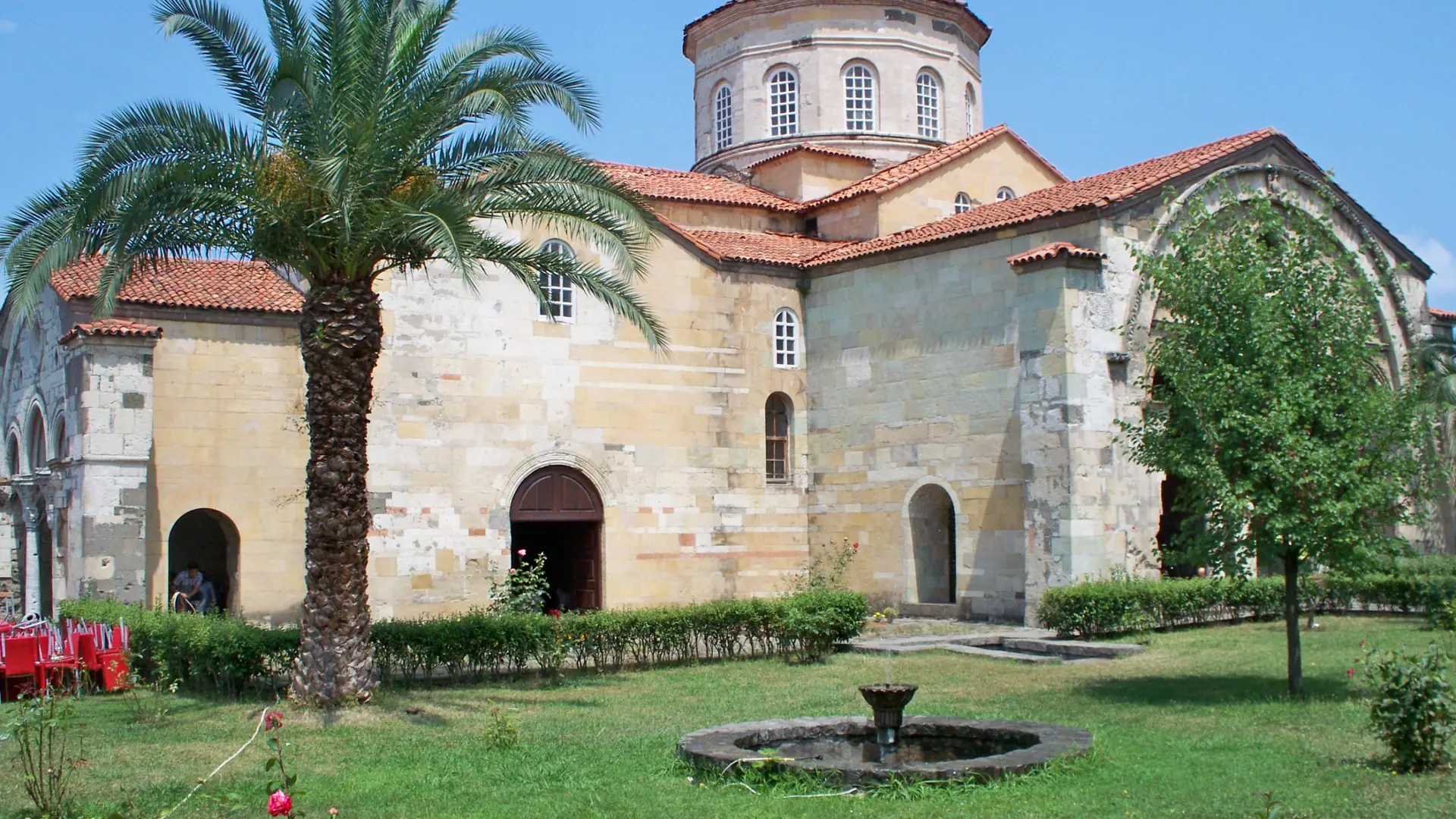 Hagia Sophia’s pale stone walls, palm and garden fountain in Trabzon, shown as a wider city scene