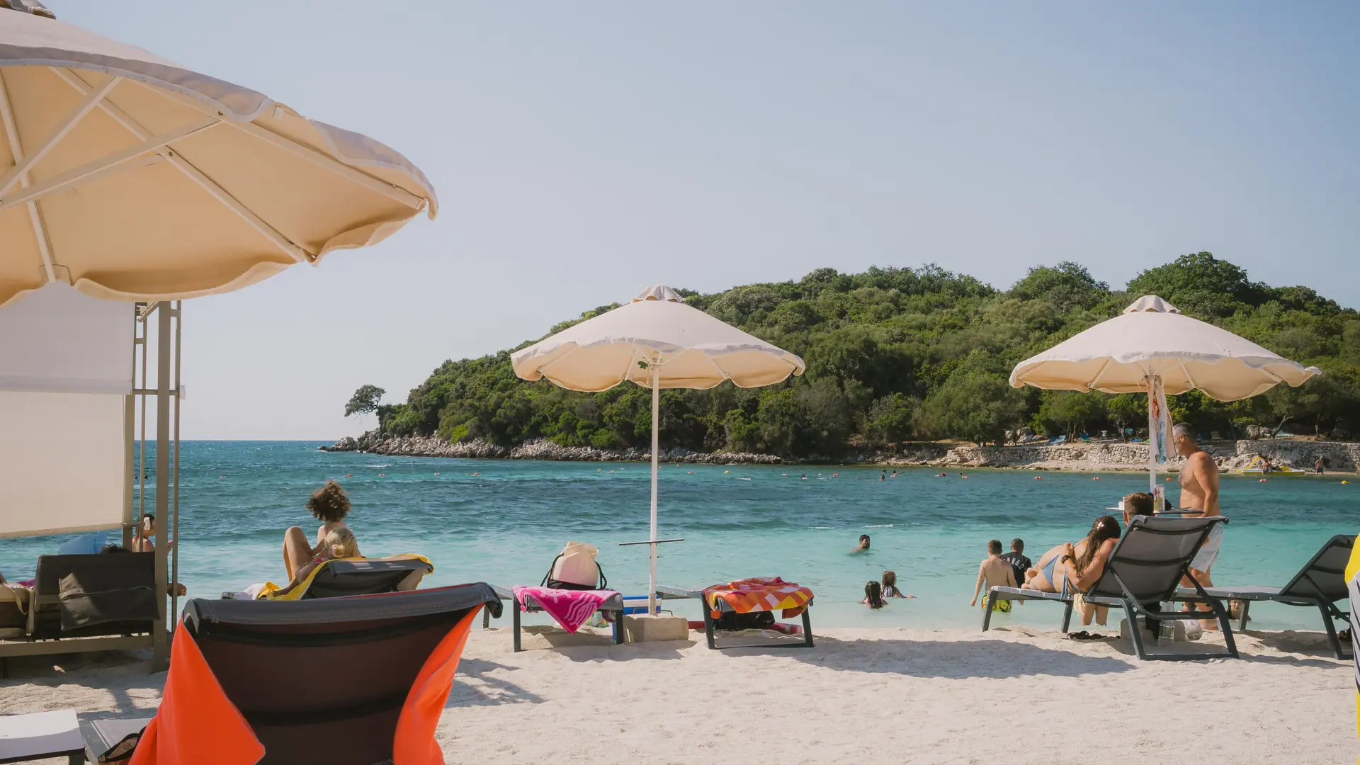 Sunbathers, umbrellas and shallow turquoise water at Ksamil's main beach, with a wooded islet behind