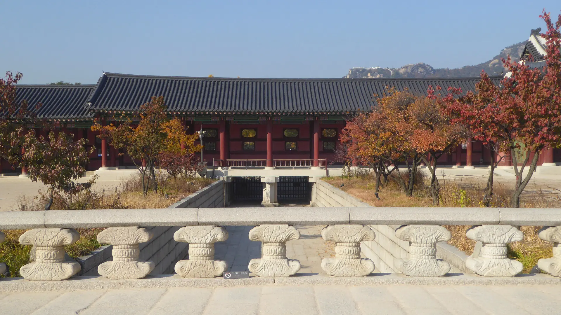 Gyeongbokgung Palace's red hanok hall and tiled roofline under a clear blue sky, with autumn trees and a mountain behind