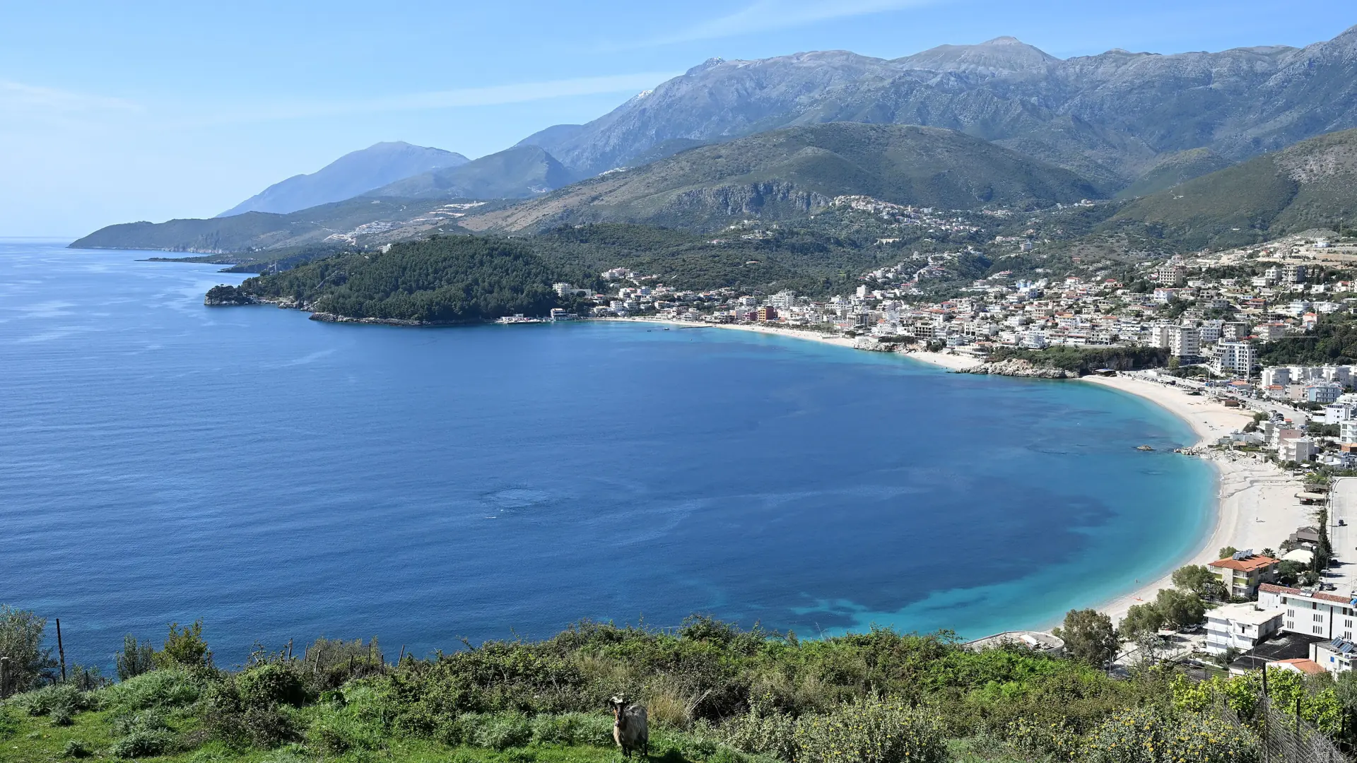 Himarë's curving bay and town below the mountains, seen from the south across clear blue water on a sunny day