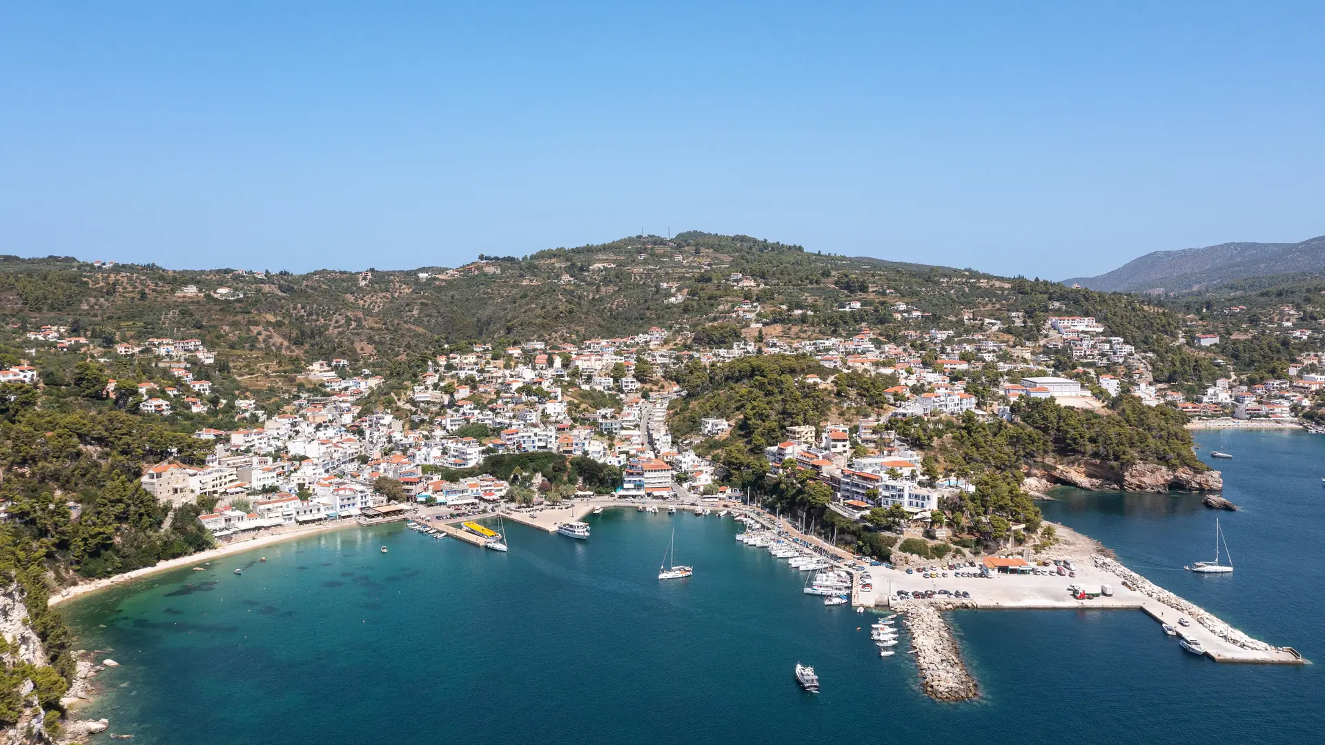 Aerial view of Patitiri harbour on Alonnisos, with a curved shore, boats and hillside houses